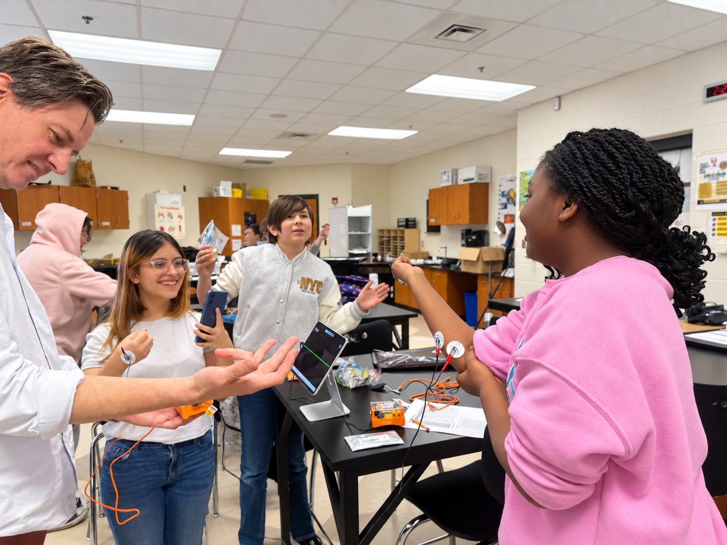 Middle school students wearing electrodes on their arms test muscle signals with Backyard Brains equipment and a tablet as a Foundation scientist holds out his hand during a Brain Explorers workshop