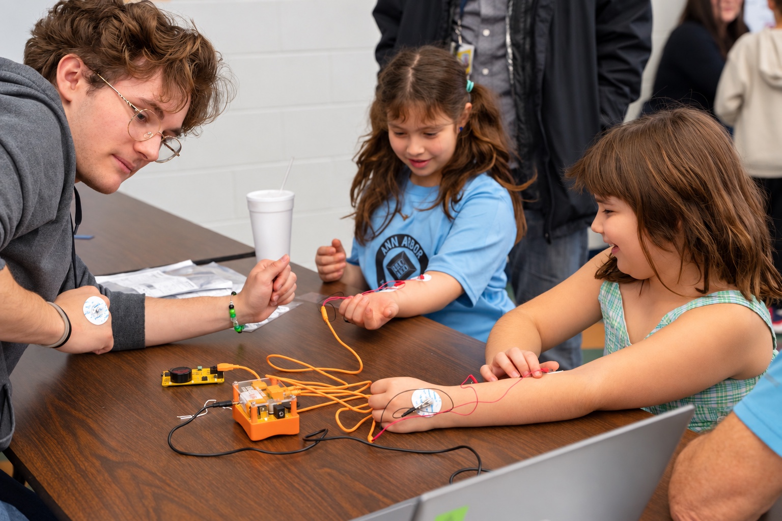 A university volunteer with an electrode on his arm guides two girls as they record their arm muscle signals with Backyard Brains equipment at Family Brain Night at Bach Elementary School