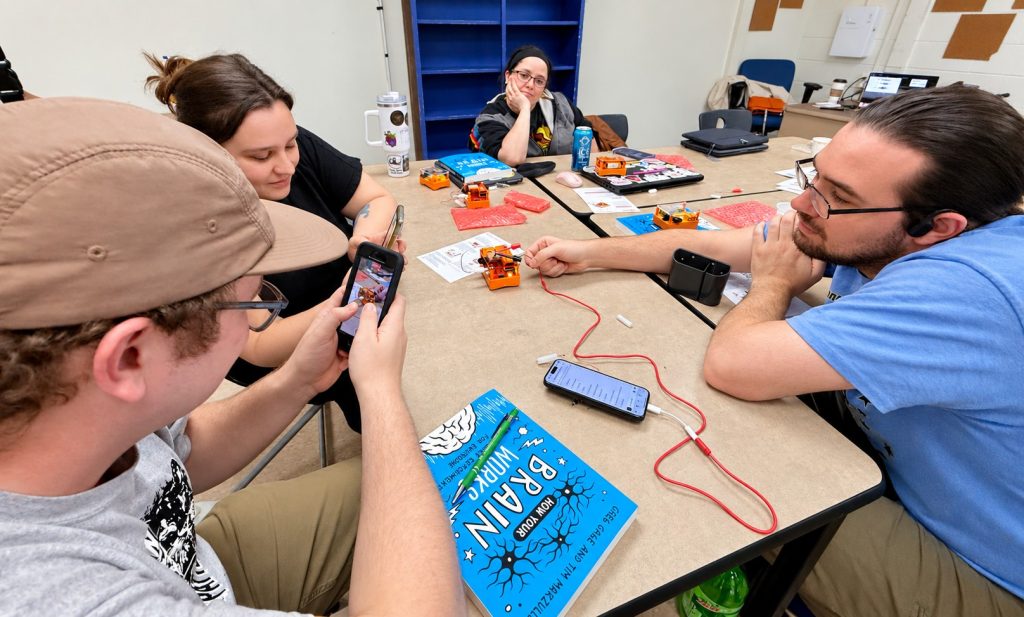 Educators around a classroom table record signals with orange Backyard Brains kits and their phones, with a copy of How Your Brain Works on the table