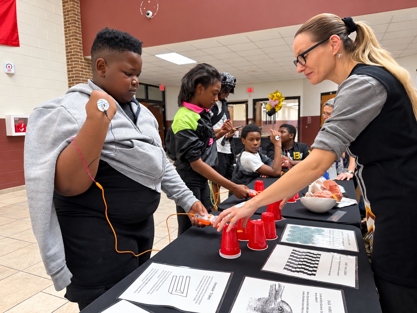 A student wearing an electrode on his arm uses his muscle signals to control a gripper that stacks red cups while an educator guides him at the Science Expo at Ypsilanti Community Middle School