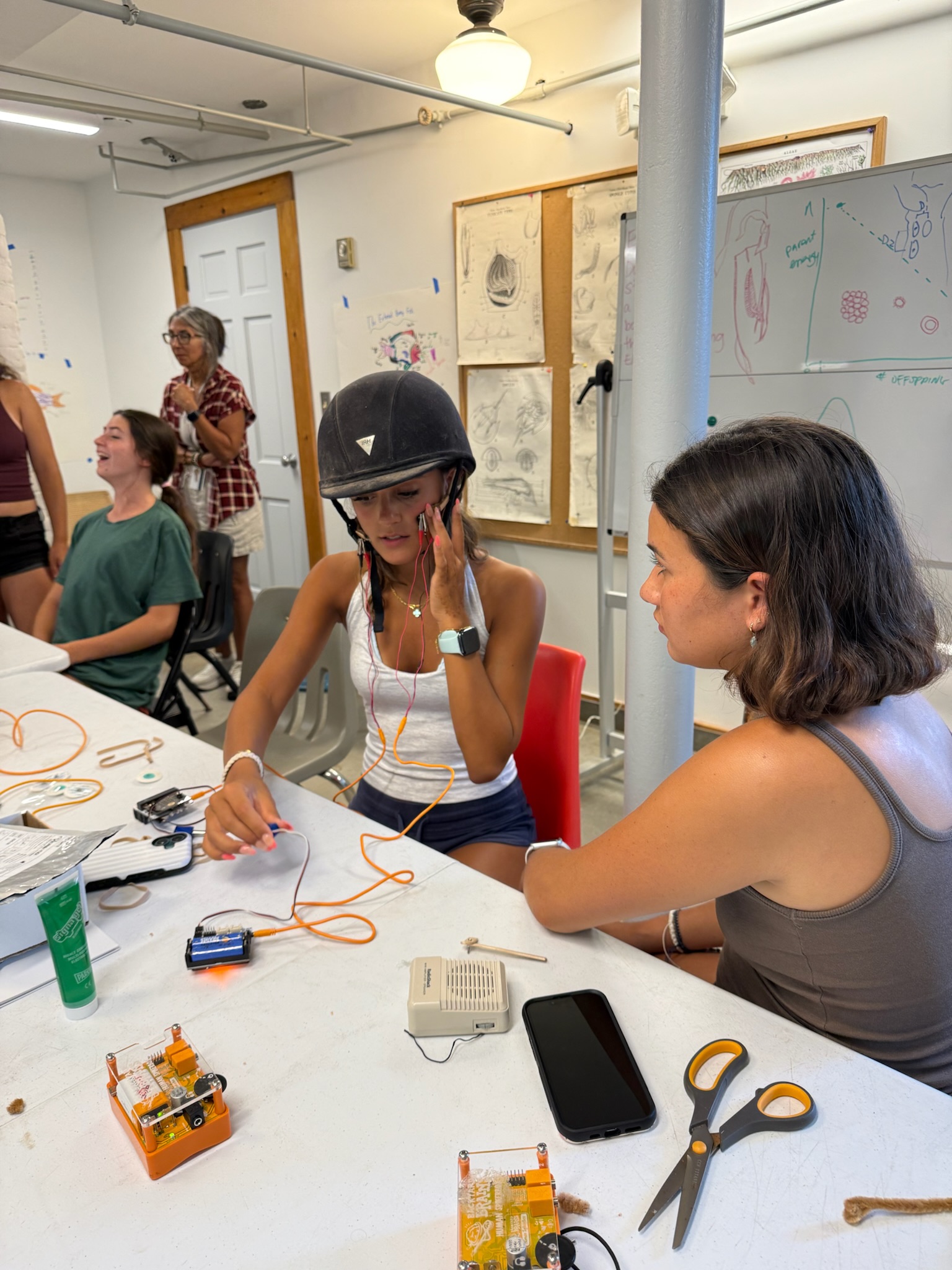 A student wearing a helmet fitted with recording electrodes works with Backyard Brains equipment while a fellow participant watches during the Summer Neuroscience Teaching Fellowship at the Children’s School of Science
